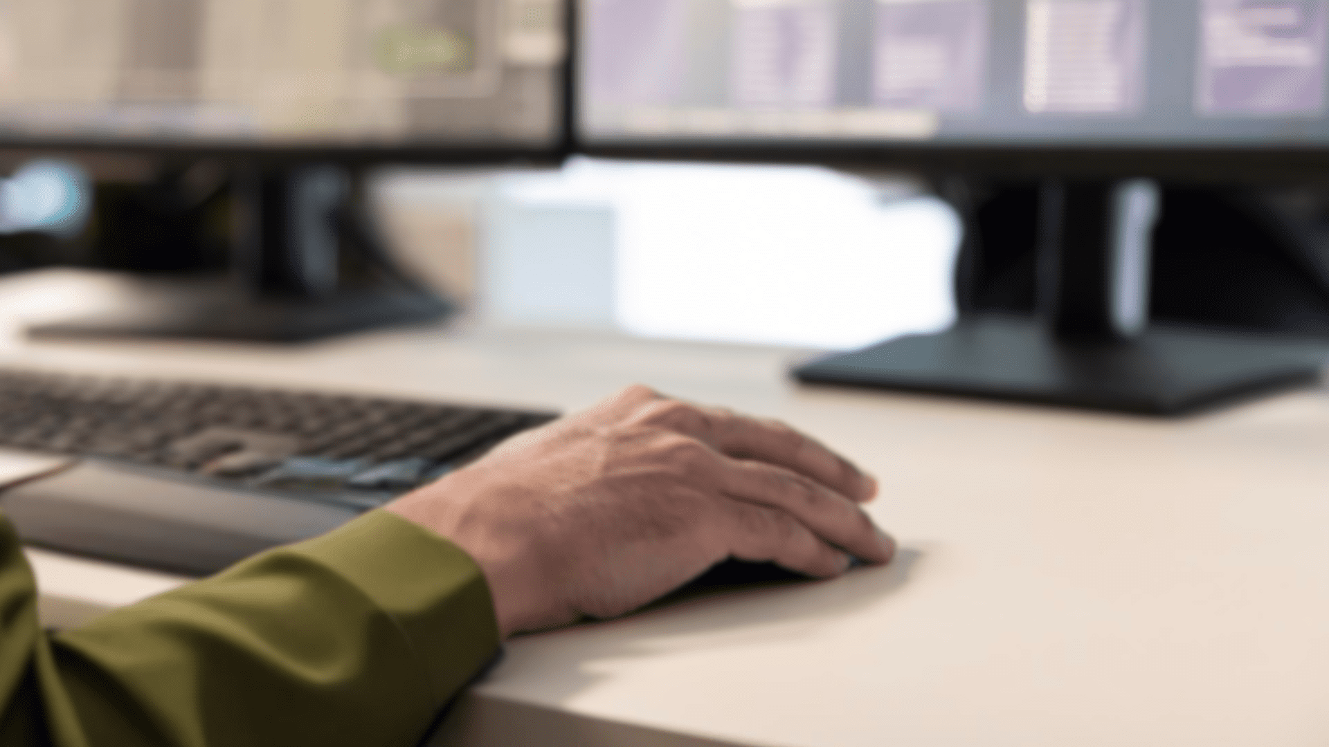 יד על עכבר מחשב מול שני מסכי עבודה ברקע עם תצוגת מידע. החולצה בצבע ירוק זית, מתכתבת עם סגנון צבאי